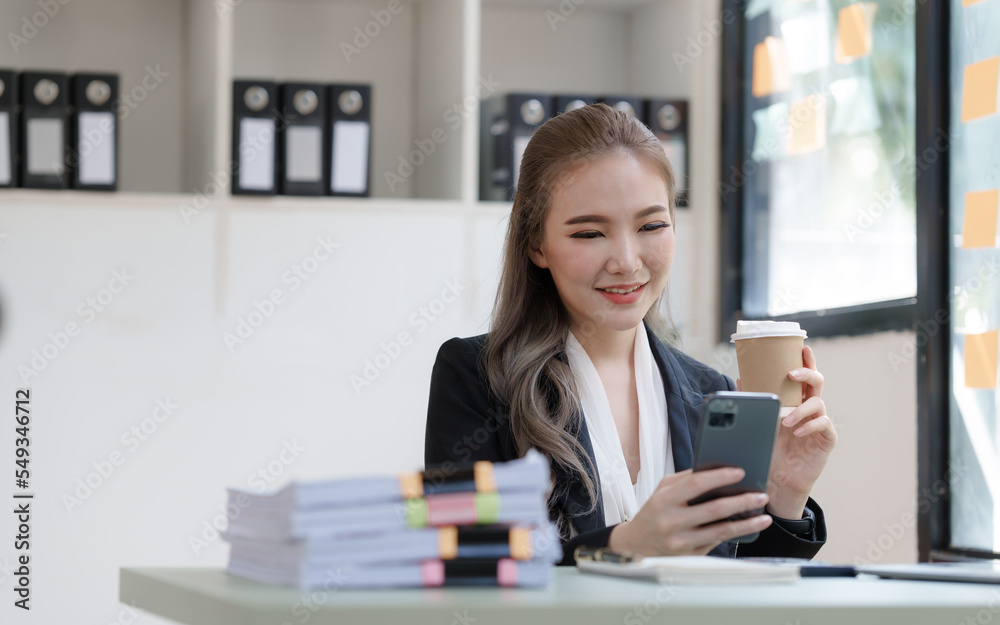 Young attractive Asian business woman with messy desk in office, sitting at the desk, working at contemporary office and documents. holding coffee cup, using mobile Young attractive Asian business woman with messy desk in office, sitting at the desk, working at contemporary office and documents. holding coffee cup, using mobile