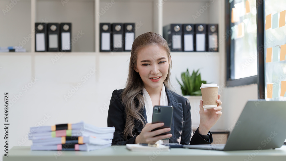Young attractive Asian business woman with messy desk in office, sitting at the desk, working at contemporary office and documents. holding coffee cup, using mobile Young attractive Asian business woman with messy desk in office, sitting at the desk, working at contemporary office and documents. holding coffee cup, using mobile