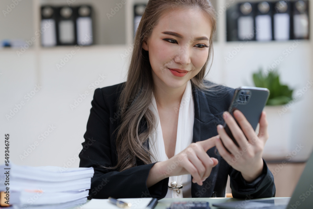 Young attractive Asian business woman with messy desk in office, sitting at the desk, working at contemporary office and documents. texting mobile Young attractive Asian business woman with messy desk in office, sitting at the desk, working at contemporary office and documents. texting mobile