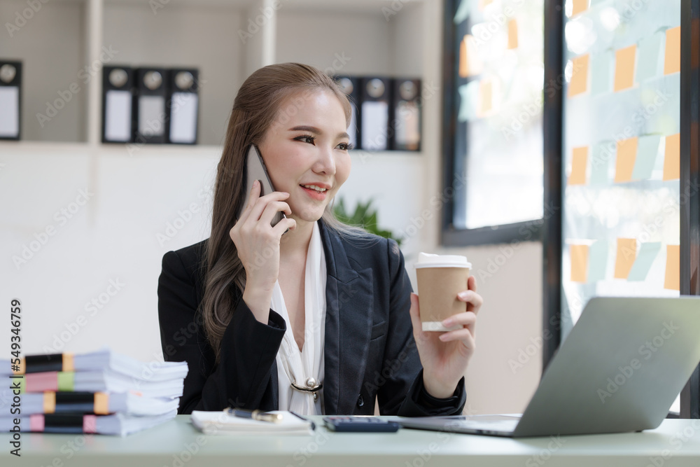 Young attractive Asian business woman with messy desk in office, sitting at the desk, working at contemporary office and documents. make a mobile phone call Young attractive Asian business woman with messy desk in office, sitting at the desk, working at contemporary office and documents. make a mobile phone call