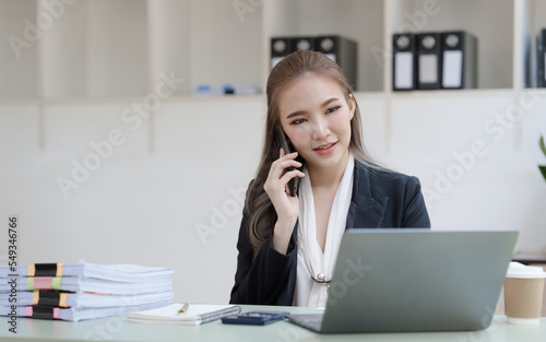 Young attractive Asian business woman with messy desk in office, sitting at the desk, working at contemporary office and documents. make a mobile phone call