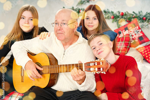 Grandfather playing guitar during christmas for grandchildren. Happy man 60s playing guitar christmas songs. Happy christmas atmosphere.