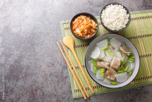 muguk light, nourishing South Korean soup combines chunks of beef and sliced Korean radishes mu served with rice and kimchi closeup on the mat on the table. Horizontal top view from above