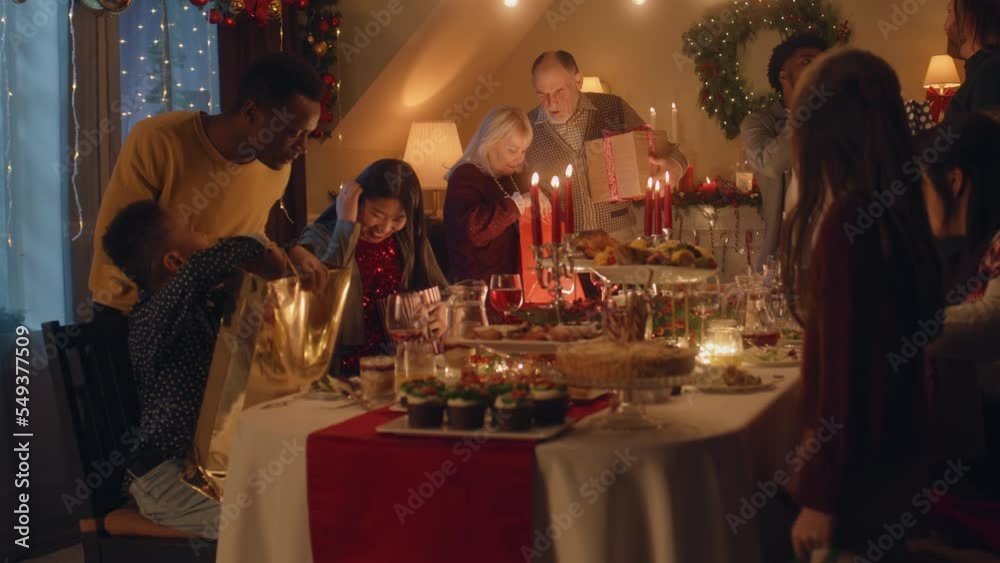 Happy multi cultural family celebrating Christmas or New Year 2023. They talking, giving and opening gifts. Served table with dishes and candles. Warm atmosphere of family Christmas dinner at home.