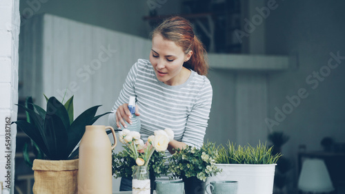 Smiling young woman is spraying green plants with water using spray bottle standing near table in modern loft style apartment. Youth, interior and household concept.