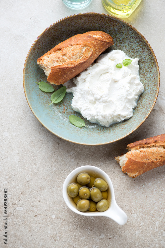 Plate with italian ricotta cheese, green olives and baguette on a beige stone background, view from above, vertical shot Plate with italian ricotta cheese, green olives and baguette on a beige stone background, view from above, vertical shot