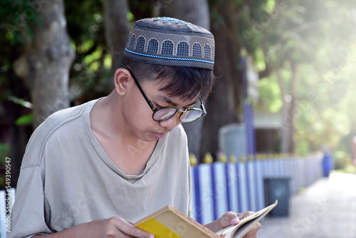 Young asian muslim boy wears eyeglasses, sitting in school park and reading his book in his free times before going back home, soft and selective focus. photo