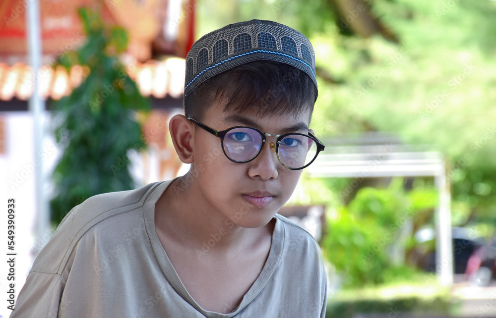 Young asian muslim boy wears eyeglasses, sitting in school park and reading his book in his free times before going back home, soft and selective focus.