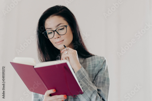Serious brunette female management consultant uses notepad for planning organization, holds pen and writes down notes or list to do, wears formal clothing, has serious expression, looks in notebook