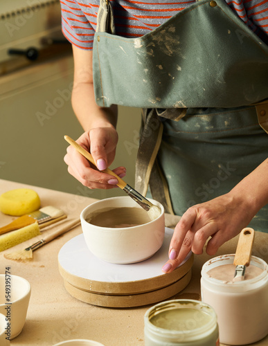 Close-up of girl painting clay mug with glaze. Woman coloring pottery in workshop with a paintbrush. Painter in green apron glazing clay pot.