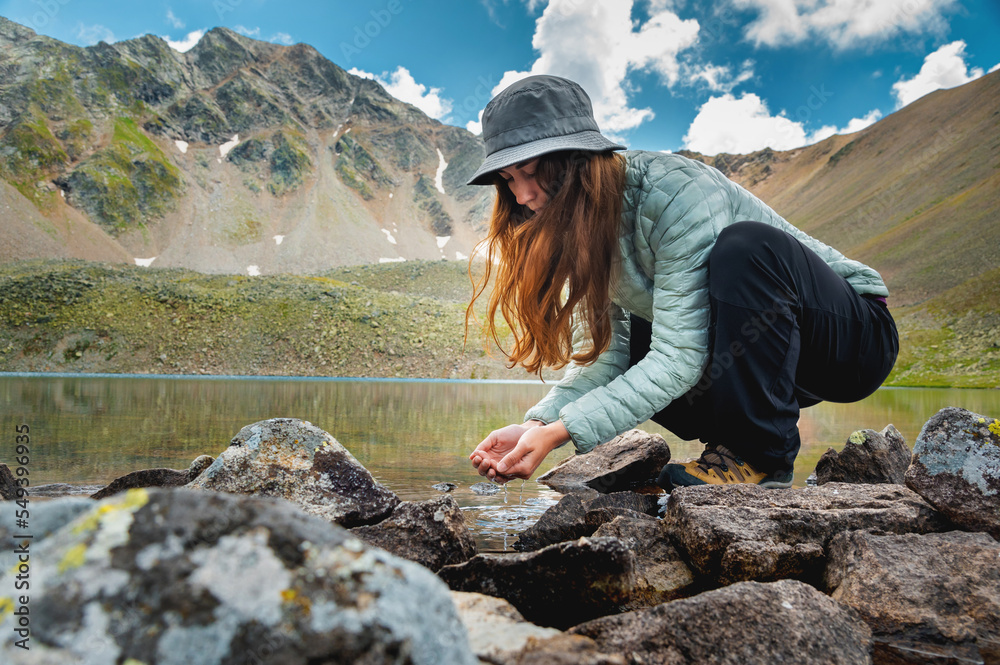 Naklejka premium young girl fills her hands with water from a stream of a cold mountain lake in order to drink and refresh herself. Happy hiker on a hike