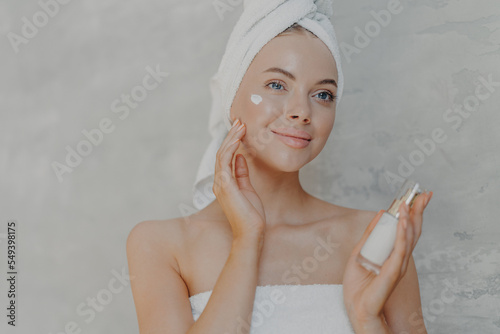 Pleased young woman has healthy glowing skin  minimal makeup  applies face lotion and looks happily into distance  wears wrapped towel on head and around body  poses against grey background.