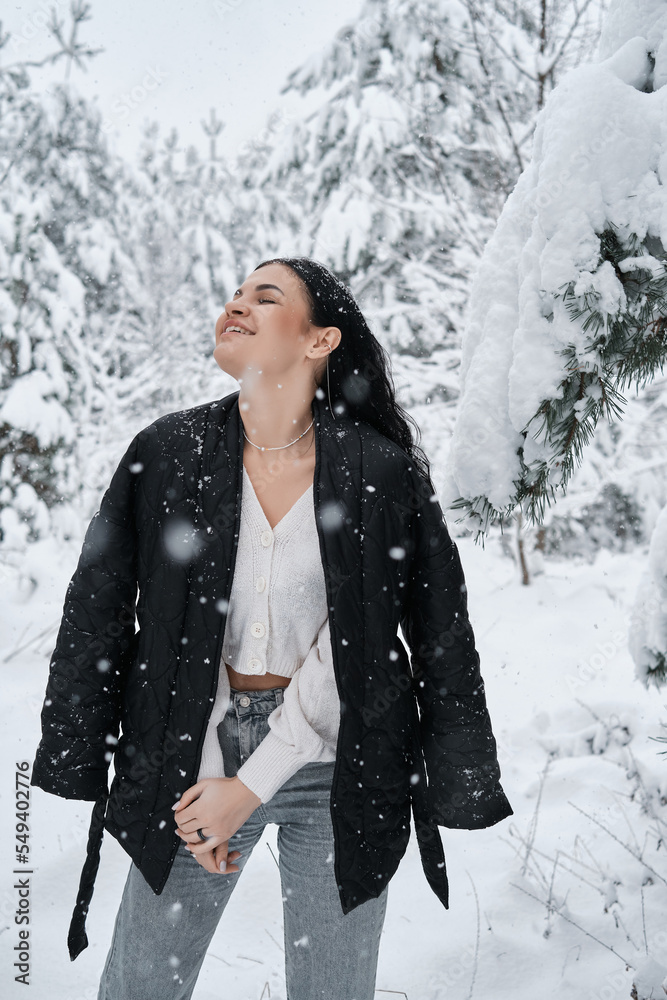 A happy girl is walking in a snowy forest. Snowfall. The girl has dark long hair. The woman is wearing a black jacket and light jeans. Christmas trees are fastened around her. A happy girl is walking in a snowy forest. Snowfall. The girl has dark long hair. The woman is wearing a black jacket and light jeans. Christmas trees are fastened around her.