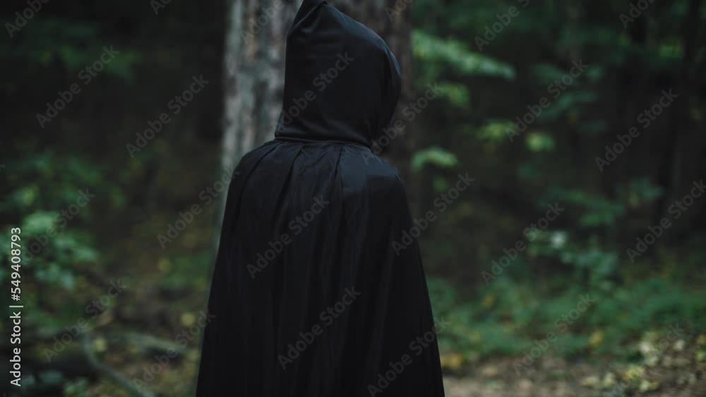 A dark figure in a black cloak with a hood walks through the enchanted forest. An unrecognizable witch from the Middle Ages walks along the path. A dark figure in a black cloak with a hood walks through the enchanted forest. An unrecognizable witch from the Middle Ages walks along the path.