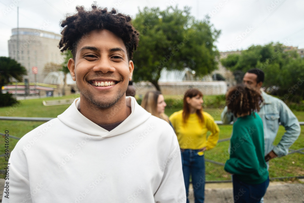 © Xavier Lorenzo - Smiling hispanic latin young man looking at camera outdoors. Portrait of african american millennial guy standing outdoors with his friends on the background. Friendship, happiness and youth concept.