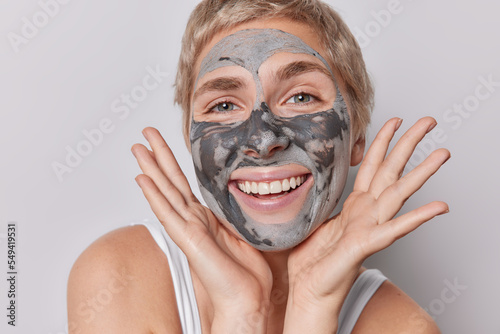 Close up shot of beautiful cheerful adult woman smiles broadly keeps hands near face shows white teeth applies beauty clay mask for skin treatment stands bare shoulders against grey background