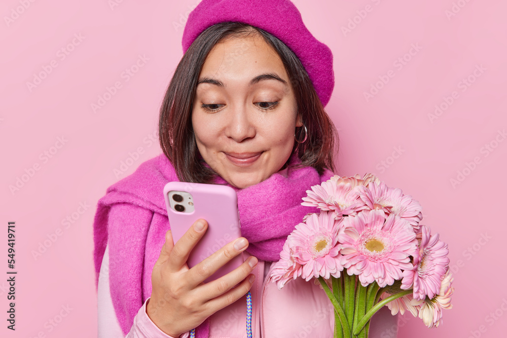Pretty Asian womman in beret and scarf around neck checks notification on cellphone holds beautiful bouquet of gerbera flowers receives on birthday isolated over pink background. Special occasion Pretty Asian womman in beret and scarf around neck checks notification on cellphone holds beautiful bouquet of gerbera flowers receives on birthday isolated over pink background. Special occasion