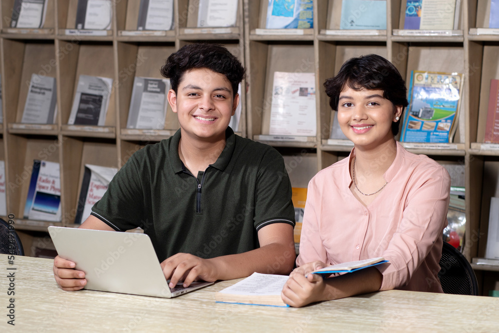 Obraz premium Young indian students reading book and using laptop studying in college library with bookshelf behind. working on assignment or project. together.