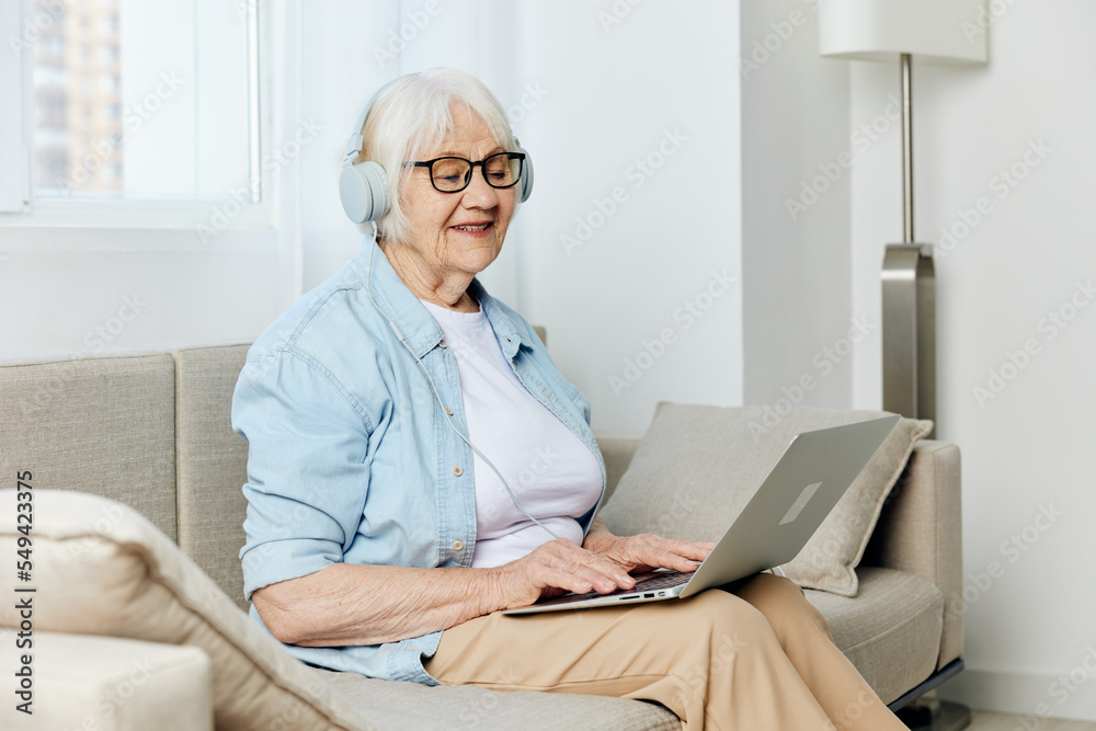 a happy elderly lady is sitting on a cozy sofa in a bright apartment, wearing stylish clothes and holding a laptop on her knees smiling joyfully with her eyes closed listening to music on headphones a happy elderly lady is sitting on a cozy sofa in a bright apartment, wearing stylish clothes and holding a laptop on her knees smiling joyfully with her eyes closed listening to music on headphones