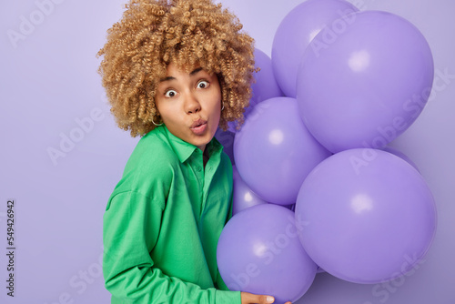 Surprised curly woman prepares for party decorates room holds bunch of inflated balloons dressed in green jumper reacts to something shocking isolated over purple background. Celebration concept