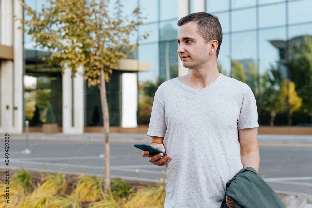 Young man holding mobile phone looking away while standing on city street Young man holding mobile phone looking away while standing on city street