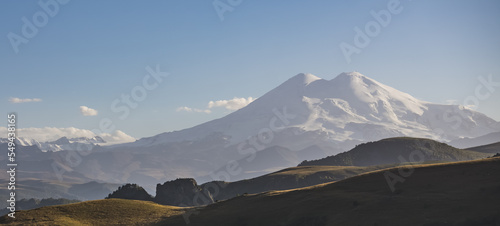 Panorama of Mount Elbrus with two peaks with snow and glaciers, grassy hills in the foreground