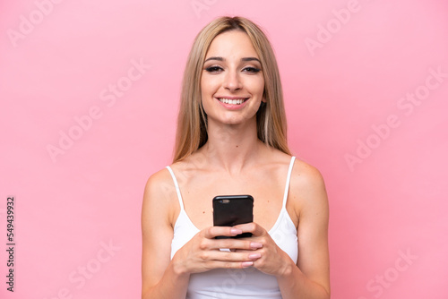 Pretty blonde woman isolated on pink background looking at the camera and smiling while using the mobile
