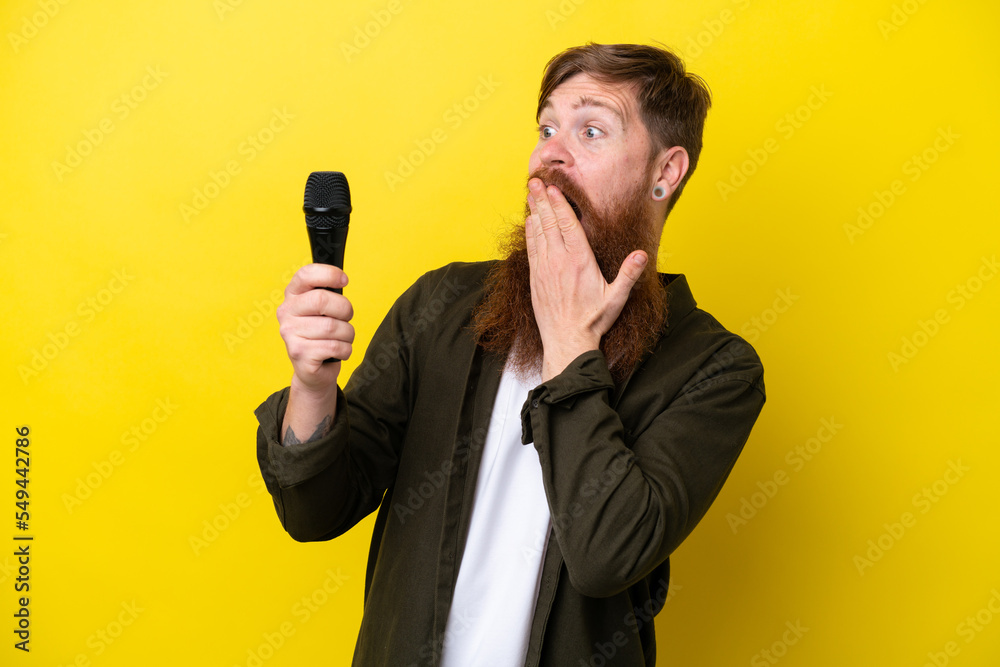 Naklejka premium Redhead man with beard picking up a microphone isolated on yellow background with surprise and shocked facial expression