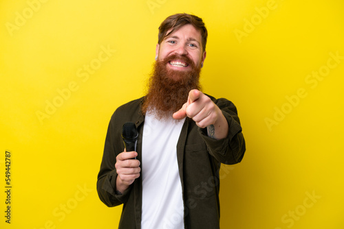 Redhead man with beard picking up a microphone isolated on yellow background points finger at you with a confident expression