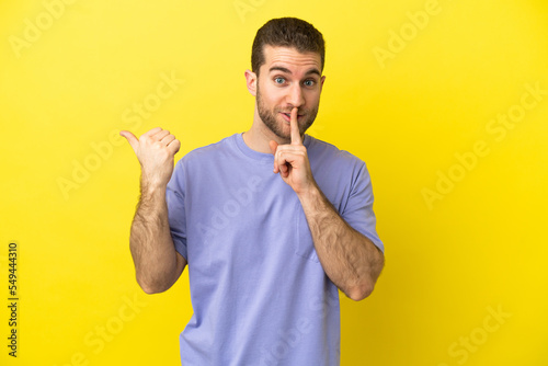 Handsome blonde man over isolated yellow background pointing to the side and doing silence gesture