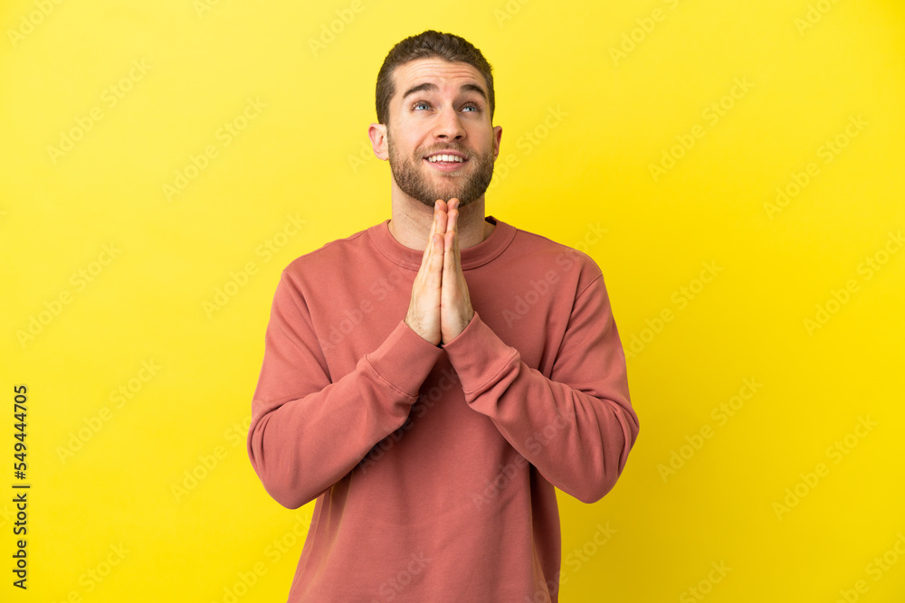 Handsome blonde man over isolated yellow background keeps palm together. Person asks for something Handsome blonde man over isolated yellow background keeps palm together. Person asks for something