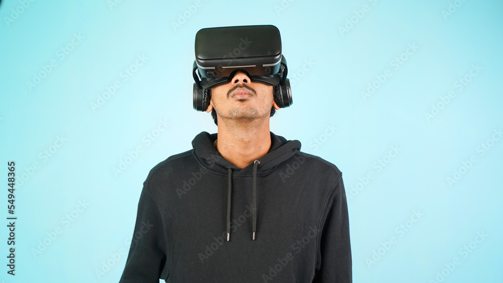 Young Indian Asian man using VR headset, glasses or goggles, isolated over blue colour indoor studio background, future, gadgets, technology, education online, studying, video game concept