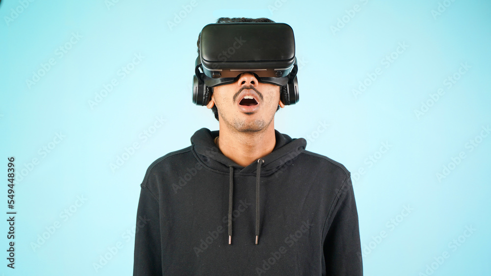 Young Indian Asian man using VR headset, glasses or goggles, isolated over blue colour indoor studio background, shocked surprised or amazed concept Young Indian Asian man using VR headset, glasses or goggles, isolated over blue colour indoor studio background, shocked surprised or amazed concept