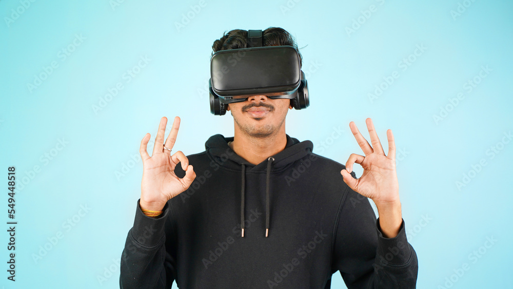 Young Indian Asian man using VR headset, glasses or goggles, isolated over blue colour indoor studio background, saying nice gesture, happy or happiness Young Indian Asian man using VR headset, glasses or goggles, isolated over blue colour indoor studio background, saying nice gesture, happy or happiness