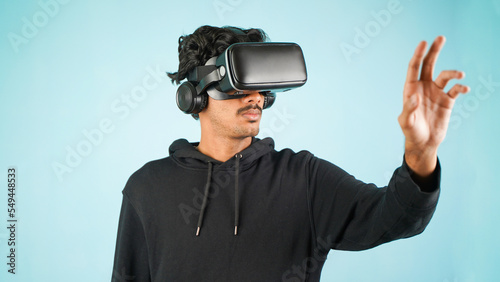 Young Indian Asian man using VR headset, glasses or goggles, isolated over blue colour indoor studio background, future, gadgets, technology, education online, studying, video game concept