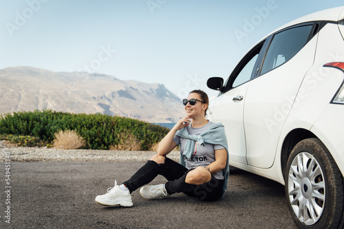 A beautiful brunette in sunglasses sits by a white car against the backdrop of the Mediterranean Sea and mountains © ruslan_shramko