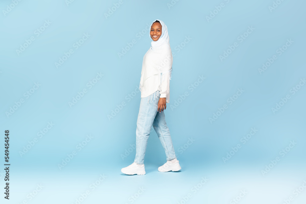 Happy african american woman in hijab walking and smiling at camera over blue studio background, full length shot Happy african american woman in hijab walking and smiling at camera over blue studio background, full length shot