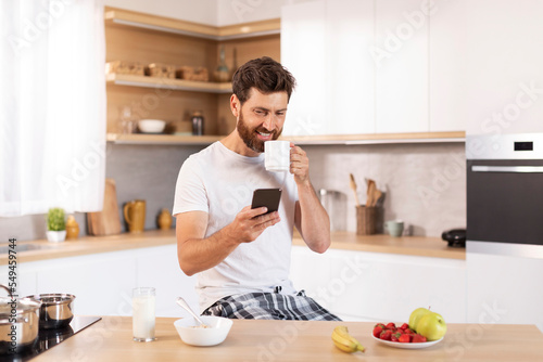 Glad mature caucasian man with beard in white t-shirt with cup drink, typing at phone in kitchen interior