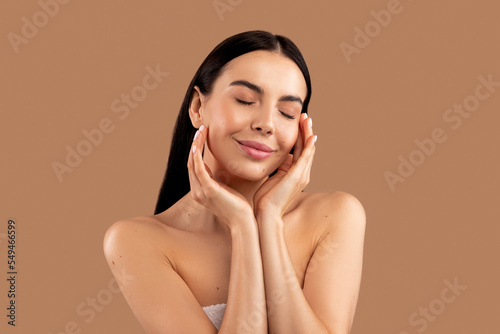 Sensual half-naked young woman touching her face, beige background