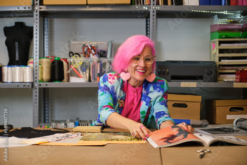 Seamstress with pink hair and colorfull clothes taking ideas from fashion magazines in a sewing workshop.
