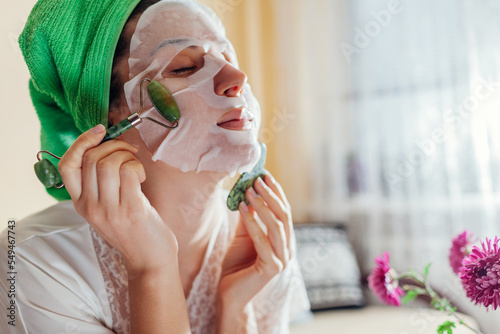 Portrait of woman with facial sheet mask applied using jade roller beauty tool and gua sha at home for unti age massage photo