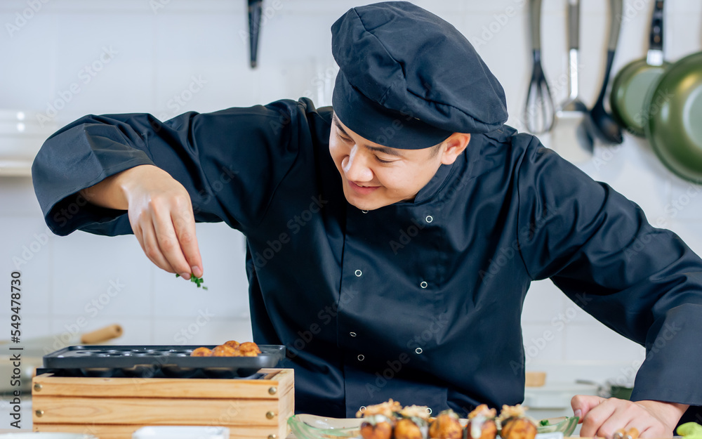 Portrait handsome professional Japanese male chef wearing black uniform, hat, cooking, making takoyaki street food, smiling with confidence, standing in kitchen. Restaurant, Hotel Concept. Portrait handsome professional Japanese male chef wearing black uniform, hat, cooking, making takoyaki street food, smiling with confidence, standing in kitchen. Restaurant, Hotel Concept.