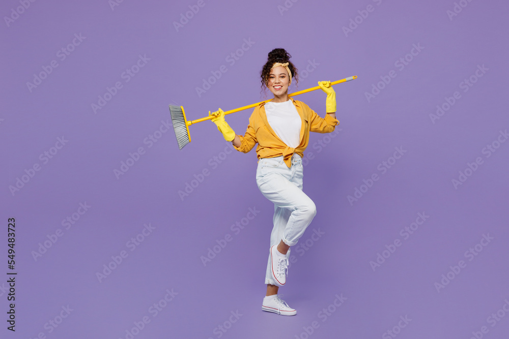 Full body side view young housekeeper woman wear yellow shirt tidy up hold broom sweeping brush behind herself clean house isolated on plain pastel light purple background studio. Housework concept. Full body side view young housekeeper woman wear yellow shirt tidy up hold broom sweeping brush behind herself clean house isolated on plain pastel light purple background studio. Housework concept.