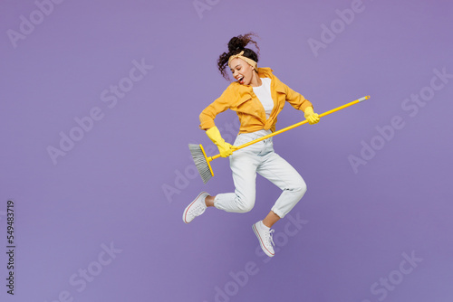 Full body side view smiling cheerful fun young housekeeper woman wear yellow shirt tidy up jump high hold broom sweep floor isolated on plain pastel light purple background studio. Housework concept.