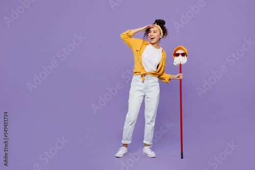 Full body young housekeeper woman wear yellow shirt tidy up hold in hand mop with hat and glasses hold head at forehead look aside isolated on plain pastel light purple background Housework concept