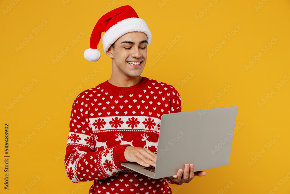 Merry student young IT man wear red knitted christmas sweater Santa hat posing hold use work on laptop pc computer isolated on plain yellow background. Happy New Year 2023 celebration holiday concept. Merry student young IT man wear red knitted christmas sweater Santa hat posing hold use work on laptop pc computer isolated on plain yellow background. Happy New Year 2023 celebration holiday concept.