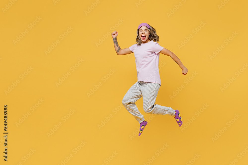 Full body young happy excited fun woman she wear purple pyjamas jam sleep eye mask rest relax at home jump high run fast hurrying isolated on plain yellow background studio portrait Night nap concept
