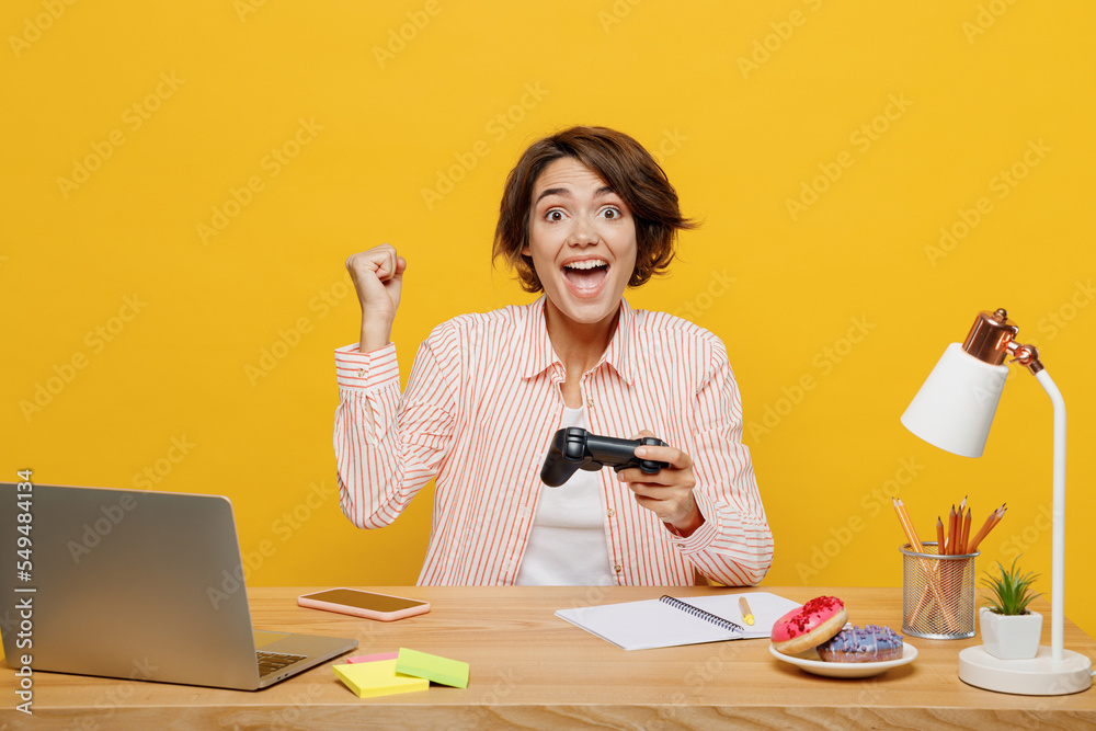 Young winner fun employee business woman wear shirt sit work at office desk with pc laptop hold play pc game with joystick console isolated on plain yellow color background Achievement career concept. Young winner fun employee business woman wear shirt sit work at office desk with pc laptop hold play pc game with joystick console isolated on plain yellow color background Achievement career concept.