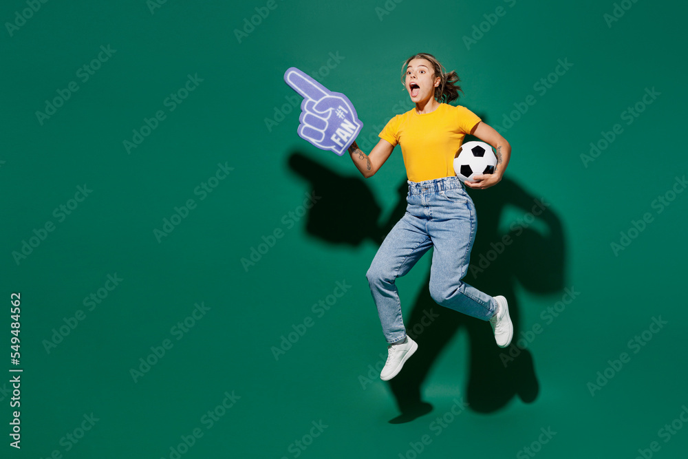 Full body young woman fan in yellow t-shirt foam 1 glove finger up point overhead cheer up support football sport team hold in hand soccer ball watch tv live stream isolated on dark green background