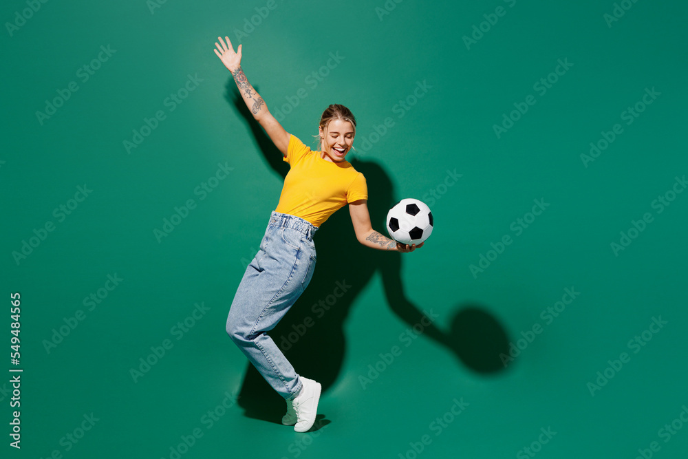 Full body fun young woman fan wear yellow t-shirt cheer up support football sport team hold soccer ball watch tv live stream stand on toes leaning back raise up hand isolated on dark green background.